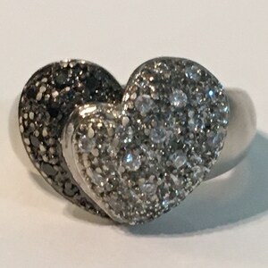 May include: A silver ring with a heart-shaped design. The heart is made of two halves, one half is set with white diamonds and the other half is set with black diamonds.