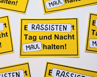 May include: Yellow rectangular stickers with black text. The stickers read "E RASSISTEN Tag und Nacht MAUL halten!" in black font. The word "MAUL" is in a white box. The stickers are scattered on a white surface.