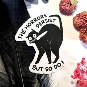Könnte beinhalten: Schwarzer Katzenaufkleber mit Messer im Maul. Der Aufkleber hat den Text "THE HORRORS PERSIST" über der Katze und "BUT SO DO I" darunter. Auf weißem Hintergrund mit getrockneten Blumen und einer schwarzen Feder.