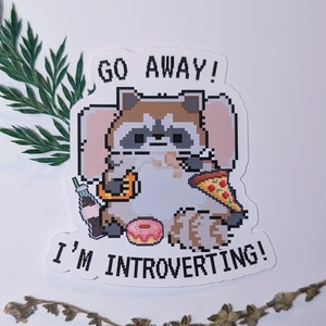 Könnte beinhalten: Ein Aufkleber mit einem pixeligen Waschbären mit einem Stück Pizza, einem Donut, einem Soda und einem Croissant. Der Waschbär sitzt auf einer rosa Decke und der Text "Go Away! I'm Introverting!" steht auf dem Aufkleber.