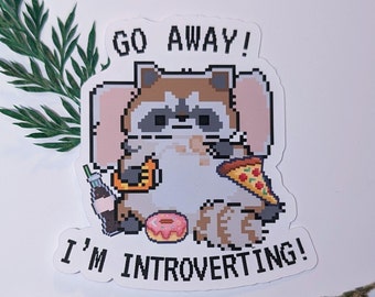 Puede incluir: Una pegatina con un mapache pixelado con una rebanada de pizza, una dona, un refresco y un croissant. El mapache está sentado en una manta rosa y el texto "Go Away! I'm Introverting!" está escrito en la pegatina.