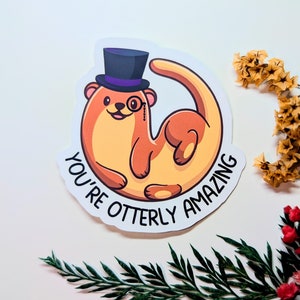 Könnte beinhalten: Ein Aufkleber mit einer Cartoon-Otter, die einen Zylinder und ein Monokel trägt. Die Otter ist zu einem Kreis zusammengerollt und der Text "You're otter-ly amazing" ist darum herum geschrieben.