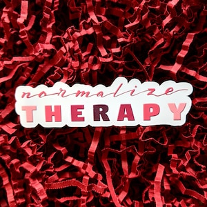 May include: A white sticker with the text "normalize therapy" in pink, red, and burgundy lettering. The sticker is on a red shredded paper background.