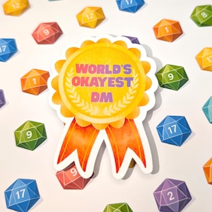 Könnte beinhalten: Ein Aufkleber mit dem Text "WORLD'S OKAYEST DM" in einem gelb-orangen Banddesign. Bunte, vielseitige Würfel mit Zahlen, darunter orange, grüne, blaue und rote Würfel, sind um den Aufkleber herum verstreut.