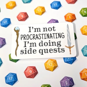 Puede incluir: Una pegatina blanca con el texto "I'm not procrastinating I'm doing side quests" e ilustraciones de un bastón y una espada. Dados poliédricos coloridos con números están dispersos.