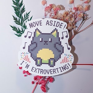 Op de afbeelding: Een witte sticker met een gepixelde kat ontwerp. De kat is grijs met een gele buik en groene voeten. De sticker zegt "Move Aside! I'm Extroverting!" met muzieknoten en feestmutsen.