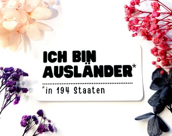 May include: White card with black text that reads "Ich bin Ausländer* in 194 Staaten". The card is surrounded by pink, red, and purple flowers.