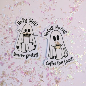 Könnte beinhalten: Zwei weiße Geisteraufkleber mit schwarzen Umrissen. Ein Geist hält eine Kamera und der Text lautet "Holy shit! You're pretty!". Der andere Geist hält eine Kaffeetasse mit dem Text "Hocus pocus Coffee for focus".
