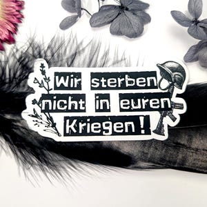 May include: A white sticker with black text that reads "Wir sterben nicht in euren Kriegen!" (We do not die in your wars!). The sticker also features a black illustration of a helmet and a rifle. The sticker is decorated with floral elements.