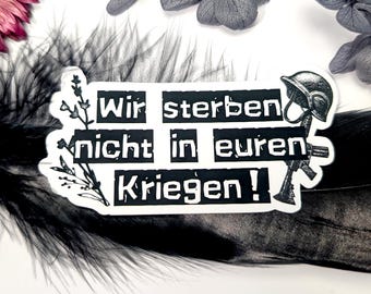 May include: A white sticker with black text that reads "Wir sterben nicht in euren Kriegen!" (We do not die in your wars!). The sticker also features a black illustration of a helmet and a rifle. The sticker is decorated with floral elements.
