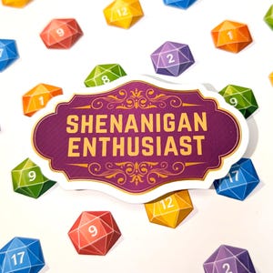 Könnte beinhalten: Ein Aufkleber mit dem Text "SHENANIGAN ENTHUSIAST" in goldfarbenen Buchstaben auf einem lila-weißen dekorativen Etikett. Um das Etikett herum sind bunte, geometrische Würfel mit Zahlen, darunter rot, blau, grün und gelb.