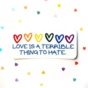 Könnte beinhalten: Ein weißer Aufkleber mit einem Regenbogen aus Herzen und dem Text "LOVE IS A TERRIBLE THING TO HATE."