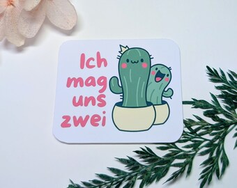 May include: A white card with a pink and green illustration of two cacti. The text on the card reads "Ich mag uns zwei".