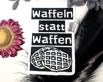 May include: A black and white sticker with the text "Waffeln statt Waffen" and an illustration of two waffles. The sticker is on a white surface, with a pink flower and a black feather in the background.