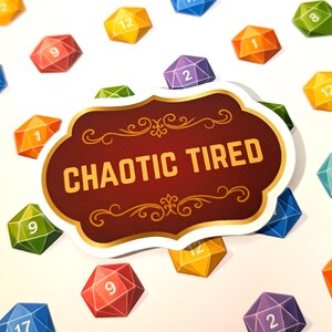 Könnte beinhalten: Ein Aufkleber mit den Worten "CHAOTIC TIRED" in goldener Schrift auf einem weinroten Hintergrund. Der Aufkleber ist von bunten, geometrischen Würfeln mit Zahlen umgeben. Die Würfel sind in verschiedenen Farben gehalten, darunter Rot, Orange, Gelb, Grün, Blau und Lila.