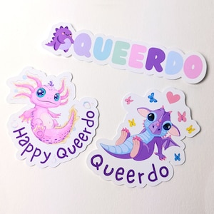 Könnte beinhalten: Drei Aufkleber mit niedlichen Comic-Tieren und dem Wort "Queerdo" in Regenbogenfarben. Der erste Aufkleber zeigt ein rosa Axolotl mit dem Text "Happy Queerdo". Der zweite Aufkleber zeigt einen lila Drachen mit dem Text "Queerdo". Der dritte Aufkleber zeigt das Wort "Queerdo" in Regenbogenfarben.