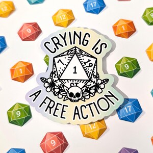 Könnte beinhalten: Ein holografischer Aufkleber mit dem Text "CRYING IS A FREE ACTION" in Schwarz. Der Aufkleber zeigt ein D20-Würfel-Design mit einem Totenkopf und floralen Akzenten. Bunte polyedrische Würfel sind um den Aufkleber verstreut.