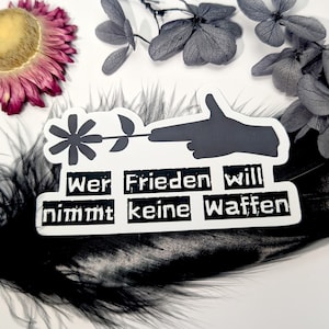 May include: A black and white sticker featuring a hand with a flower in place of a gun barrel. The sticker has the German text "Wer Frieden will nimmt keine Waffen" which translates to "Who wants peace does not take weapons". The sticker is on a black feather.
