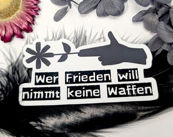 May include: A black and white sticker featuring a hand with a flower in place of a gun barrel. The sticker has the German text "Wer Frieden will nimmt keine Waffen" which translates to "Who wants peace does not take weapons". The sticker is on a black feather.