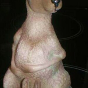 May include: A brown and tan ceramic figurine of a kangaroo with a joey in its pouch. The kangaroo is standing on all fours with its head turned to the side. The joey is peeking out of the pouch.