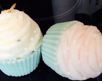 May include: Two cupcake-shaped candles, one is white with blue sprinkles and a light blue base, the other is pink with a light blue base.