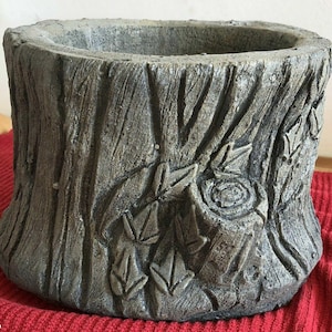May include: A gray concrete planter shaped like a tree stump with detailed carvings of leaves and a ring around the center.