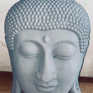 May include: A gray stone sculpture of a Buddha head with closed eyes. The head is detailed with a textured surface and a small dot on the forehead.