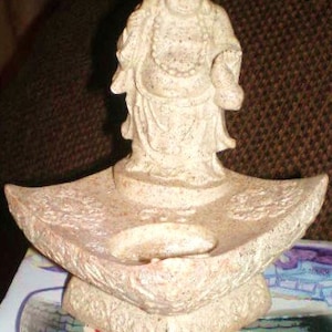 May include: A ceramic incense burner with a beige finish. The burner features a small, round bowl in the center and a decorative design around the edges. A small statue of a smiling Buddha sits atop the burner.