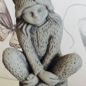 Puede incluir: Una figurita de gnomo de cerámica gris sentada en un tocón de árbol. El gnomo lleva un gorro puntiagudo y tiene una barba larga. El gnomo está sentado con las piernas cruzadas y las manos juntas en su regazo.