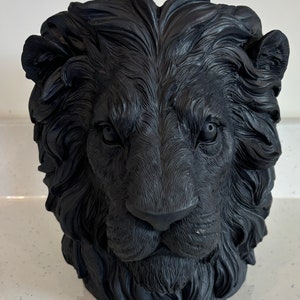 Puede incluir: Maceta de cabeza de león negra hecha de cerámica. La maceta es una representación realista de la cabeza de un león con una melena detallada.