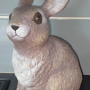 Puede incluir: Una figurita de conejo gris con ojos marrones y una mancha blanca en el pecho. El conejo está sentado con las orejas levantadas.