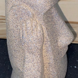 May include: A speckled, tan-coloured ceramic vase in the shape of a Moai statue from Easter Island. The vase has a detailed face with a prominent nose, closed eyes, and hands resting on the chest. The vase has a cylindrical opening at the top.