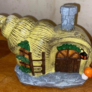 May include: A yellow and brown ceramic snail house with a gray chimney, a green vine, a brown door, and a mushroom on the side.