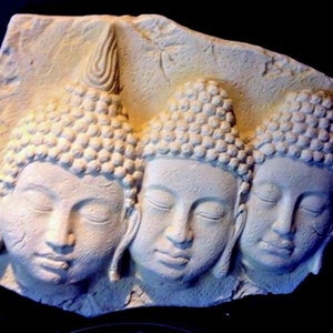 May include: White stone relief sculpture of three Buddha faces with closed eyes. The sculpture is carved in a rectangular shape and has a rough texture.