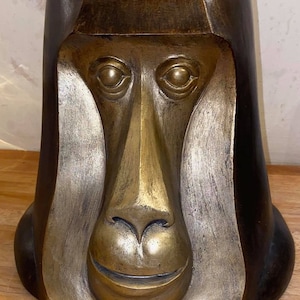 May include: A bronze sculpture of a monkey's face. The sculpture is in the shape of a vase, with the monkey's face forming the opening. The sculpture is a dark bronze colour with a gold patina.