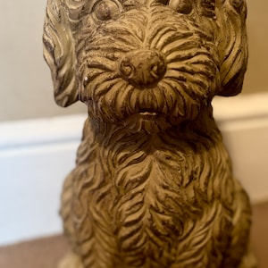 May include: A small, brown, ceramic dog statue with a shaggy, textured coat. The dog is sitting with its head tilted slightly to the side.