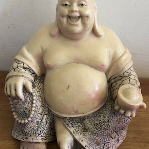 May include: A smiling, white ceramic statue of the Laughing Buddha, sitting with a beaded necklace in one hand and a round object in the other. The statue is decorated with a mosaic-like pattern.
