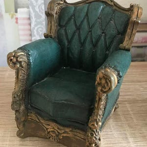 May include: A miniature armchair with a green velvet upholstery and gold-painted ornate details. The chair has a high back and a decorative crest.