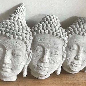 May include: Three grey Buddha head sculptures with closed eyes and serene expressions. The heads are arranged side-by-side, each with a textured surface and detailed hair. The sculptures are likely made of stone or a similar material.