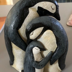 May include: A ceramic penguin family figurine. The three penguins are painted in black and white with a textured finish. The largest penguin is standing upright with its wings wrapped around the middle penguin. The middle penguin is standing upright with its wings wrapped around the smallest penguin. The smallest penguin is standing upright with its wings tucked in.