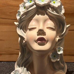 May include: A white ceramic bust of a woman with closed eyes and a flower crown. The flowers are blue with green centres. The bust is a decorative piece.