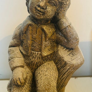 Puede incluir: Una escultura de gnomo de jardín de piedra gris sentada en un tocón de árbol. El gnomo lleva un sombrero y tiene una expresión pensativa en su rostro.