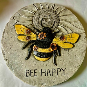 May include: A gray stone garden stepping stone with a 3D bee design. The bee is black and yellow with brown and yellow flowers. The stone has the text "BEE HAPPY" written on it.
