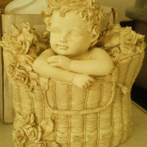 Może przedstawiać: Biała ceramiczna figurka cheruba siedzącego w wiklinowym koszyku z kwiatowymi akcentami. Cherub ma kręcone włosy i trzyma kosz obiema rękami.