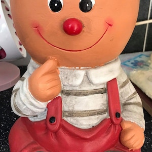 Peut inclure: Un pot en terre cuite en forme de personne souriante portant une salopette rouge et une chemise à rayures blanches et brunes. Le pot a un nez rouge et des yeux noirs.