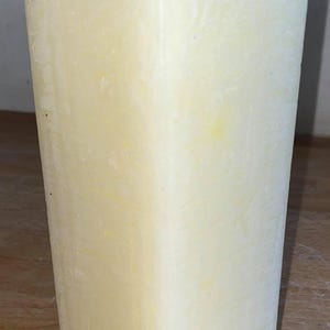 May include: A large, off-white pillar candle with a single wick. The candle has a textured surface and is rectangular in shape.