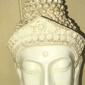 Può includere: Scultura in ceramica bianca di testa di Buddha con una corona di fiori e un nodo decorativo. La scultura ha gli occhi chiusi e un'espressione serena.
