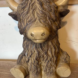 Puede incluir: Figura detallada de una vaca de las Highlands sentada. La escultura presenta una cara y pezuñas de color marrón claro, con pelo largo y lanudo de color marrón. La vaca tiene cuernos grandes y curvados y está en una posición relajada.