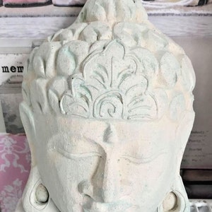 Può includere: Una scultura in gesso dipinta di bianco e verde di una testa di Buddha con un motivo floreale sulla fronte.