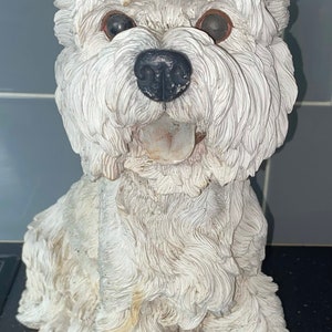 Może przedstawiać: Biała ceramiczna figurka psa rasy West Highland White Terrier siedzącego z wystawionym językiem. Pies ma brązowe oczy i czarny nos.